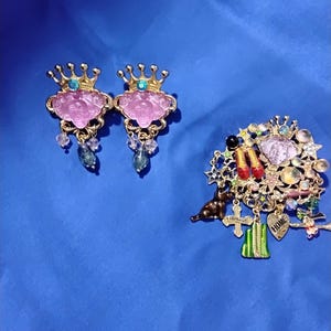Könnte beinhalten: Zwei goldfarbene kronenförmige Ohrringe mit geschnitzten rosa Gesichtern, blauen Edelsteinen und hängenden Kristallen sowie eine goldfarbene Brosche mit verschiedenen Anhängern, darunter rote Schuhe, ein grünes Gebäude, ein Herz mit der Aufschrift HOME und ein schwarzer Hund.