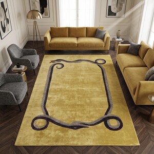 May include: A rectangular area rug in a mustard yellow hue featuring a dark grey border design composed of two intertwined snakes. The snakes form a continuous frame around the centre of the rug, with their heads meeting at the top and bottom.