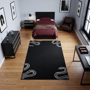 May include: A rectangular area rug featuring a solid black background with white line art illustrations of snakes positioned in each of the four corners. The rug is placed on a light-coloured wooden floor in a room with black furniture and decor.