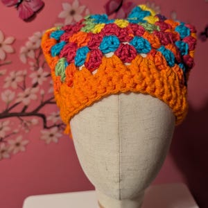 May include: A handmade crochet hat in vibrant colors. The hat features a bright orange brim and a crown of colorful squares in shades of red, blue, yellow, and green. The hat is displayed on a mannequin head.