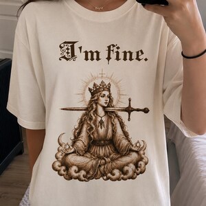 Puede incluir: Camiseta color crema con el texto "I'm fine." sobre una ilustraci&oacute;n sepia de una mujer coronada con una espada en la boca, sentada en una nube. La mujer est&aacute; rodeada por un halo.