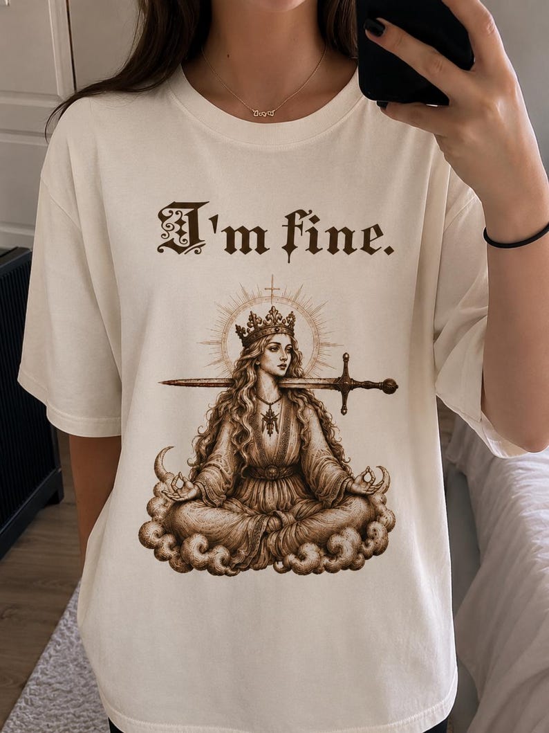 Puede incluir: Camiseta color crema con el texto "I'm fine." sobre una ilustraci&oacute;n sepia de una mujer coronada con una espada en la boca, sentada en una nube. La mujer est&aacute; rodeada por un halo.