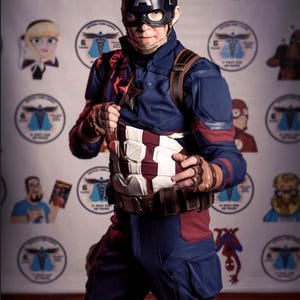 May include: A person in a Captain America costume, featuring a navy blue and red suit with a white and red striped chest piece. The helmet has a silver "A". The background has cartoon character images.