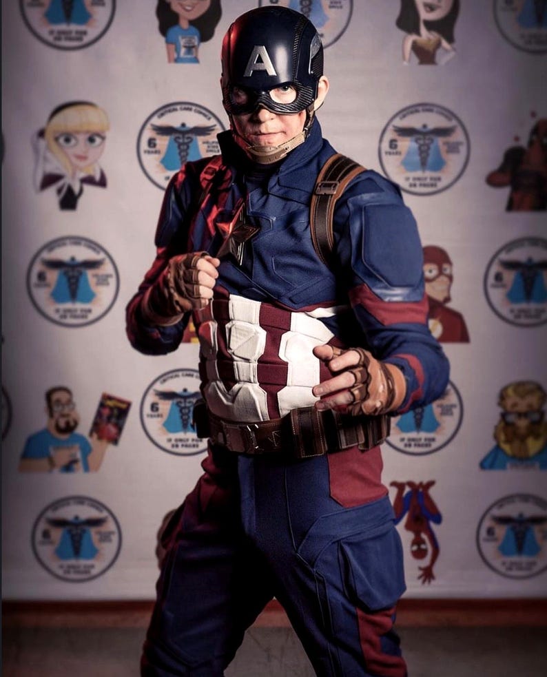 May include: A person in a Captain America costume, featuring a navy blue and red suit with a white and red striped chest piece. The helmet has a silver "A". The background has cartoon character images.