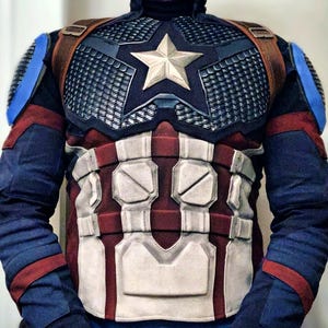 May include: A Captain America costume featuring a navy blue and red color scheme. The chest piece has a silver star and a grid-like pattern. Brown straps cross the shoulders. The costume includes blue sleeves with red accents.