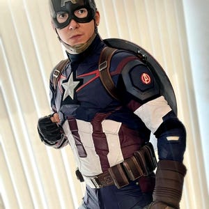 May include: Captain America costume featuring a navy blue and white suit with a star emblem, a black helmet with the letter "A", and a shield. The costume includes a brown belt, gloves, and a shield. The overall design is a superhero theme.