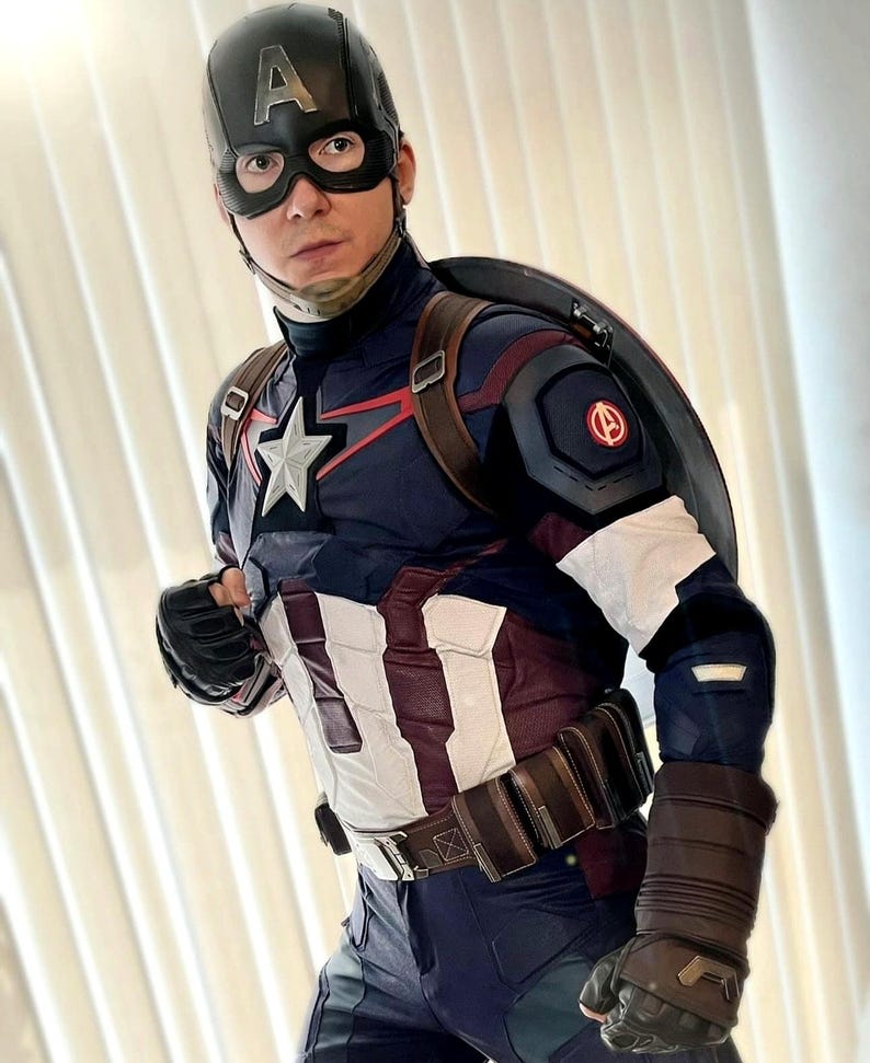May include: Captain America costume featuring a navy blue and white suit with a star emblem, a black helmet with the letter "A", and a shield. The costume includes a brown belt, gloves, and a shield. The overall design is a superhero theme.