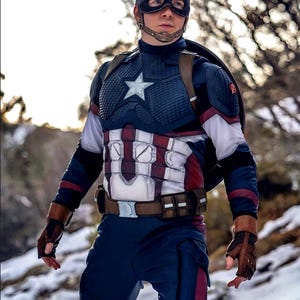 May include: A Captain America costume featuring a navy blue helmet with a white "A", a star-emblazoned chest piece, and a brown belt. The costume includes a shield, gloves, and a blue and red color scheme. The background is a snowy, outdoor setting.