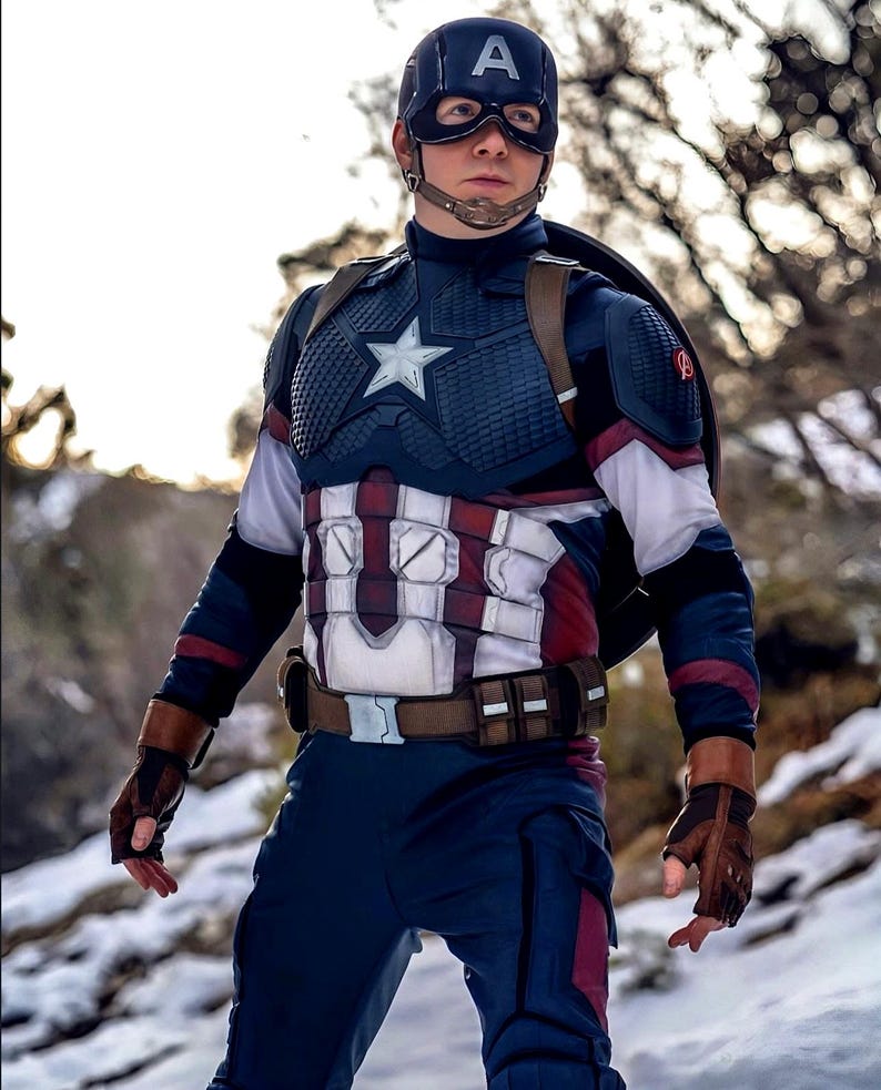 May include: A Captain America costume featuring a navy blue helmet with a white "A", a star-emblazoned chest piece, and a brown belt. The costume includes a shield, gloves, and a blue and red color scheme. The background is a snowy, outdoor setting.