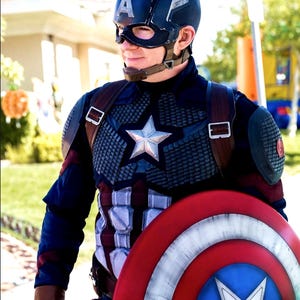 May include: A person in a Captain America costume, featuring a navy blue helmet with a white "A", a star-emblazoned chest plate, and a red, white, and blue shield. The costume includes brown leather straps and gloves.