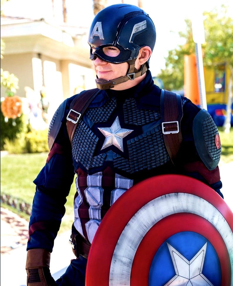 May include: A person in a Captain America costume, featuring a navy blue helmet with a white "A", a star-emblazoned chest plate, and a red, white, and blue shield. The costume includes brown leather straps and gloves.