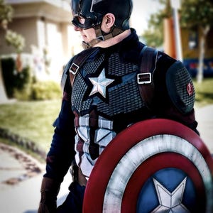 May include: A Captain America costume featuring a navy blue helmet, a star-emblazoned chest plate, and a red, white, and blue shield. The costume includes a star on the chest and a matching star on the shield. The overall design is a patriotic superhero theme.