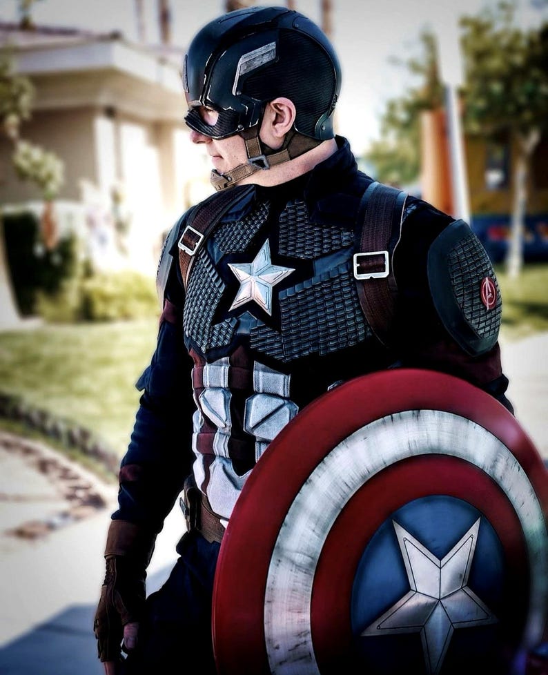 May include: A Captain America costume featuring a navy blue helmet, a star-emblazoned chest plate, and a red, white, and blue shield. The costume includes a star on the chest and a matching star on the shield. The overall design is a patriotic superhero theme.