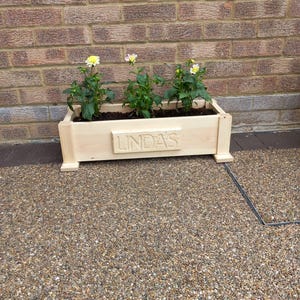 May include: A rectangular light-coloured wooden planter box featuring the name LINDA'S carved into a central raised panel. The planter contains three green plants with small yellow and white flowers, supported by four small feet at the base.