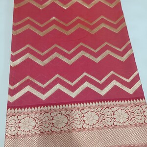May include: A vibrant red fabric featuring a repeating horizontal gold chevron pattern across the main body. The lower section displays a wide, intricate border with detailed floral motifs and a fine cross-hatched texture in metallic gold.