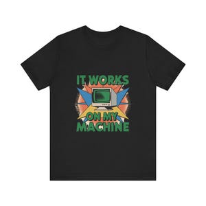 Peut inclure: Un t-shirt noir à manches courtes avec une impression graphique d'un écran d'ordinateur vintage centré au-dessus du texte IT WORKS ON MY MACHINE en lettres vertes, sur un fond de formes géométriques en étoile bleues et orange.