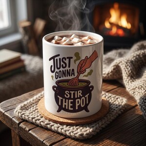 May include: A white ceramic mug filled with a hot beverage topped with marshmallows. The mug features a graphic of a hand stirring a dark purple cauldron with a wooden spoon and the text "JUST GONNA STIR THE POT" in dark purple and green lettering.