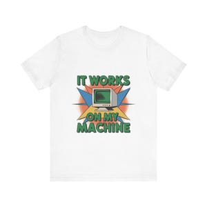 May include: A white short-sleeve t-shirt featuring a graphic of a vintage computer monitor centered on a starburst background. The text IT WORKS ON MY MACHINE is printed in green block letters above and below the computer illustration.