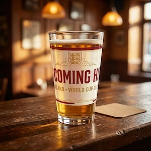 May include: A pint glass filled with beer, featuring a label with the text "COMING HOME" in red, along with the England crest. The glass sits on a wooden bar with a coaster.