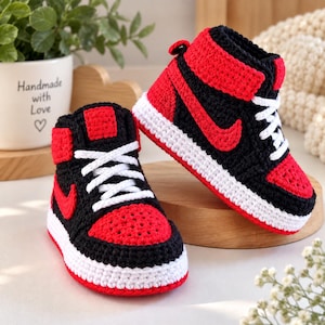 May include: A pair of high-top baby booties crocheted in a black, red, and white color scheme. The booties feature a black base with red toe boxes, red side panels, and white soles. White laces are threaded through the front of each bootie.