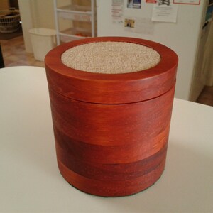 May include: A round wooden box with a fabric-covered lid. The box is a rich reddish-brown colour and has a natural wood grain.