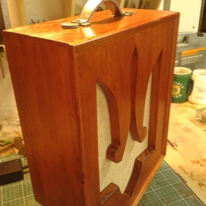 May include: A wooden box with a metal handle and a stylised trident design on the front. The box is stained a rich brown colour and has a light brown fabric covering the trident design.