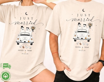 Puede incluir: Camiseta color marfil con un gráfico de dos fantasmas en un coche con el texto "JUST married", "OLIVIA & NOAH" y "10.25.26". El diseño incluye una luna creciente, estrellas, un ramo y latas arrastrándose detrás del vehículo.