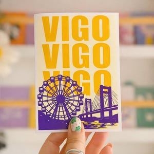 Könnte beinhalten: Ein rechteckiger Aufkleber mit dem Wort "VIGO" in gelben Blockbuchstaben. Darunter eine lila-gelbe Illustration eines Riesenrads und einer Brücke. Der Aufkleber wird von einer Hand gehalten.