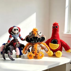 Könnte beinhalten: Drei Plüschtiere werden auf einer weißen Oberfläche präsentiert. Zu den Spielzeugen gehören eine rot-blaue spinnenartige Kreatur, ein braun-oranges Monster und eine rot-vogelförmige Figur mit gelbem Schnabel. Die Spielzeuge haben übertriebene Merkmale.