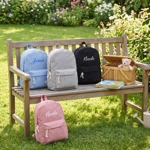 May include: Four corduroy backpacks in light blue, gray, black, and pink, each featuring a front pocket with white rope zipper pulls and a personalized name embroidered in white script. A small teddy bear sits inside a woven wooden basket.