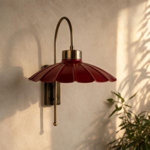 Könnte beinhalten: Eine Wandleuchte mit einem tiefroten, gewellten Metallschirm und einem geschwungenen Messingarm. Die Leuchte verfügt über eine rechteckige Messing-Wandhalterung und eine vertikale Messingstange, die unter dem Schirm in einem kleinen Kugelknopf endet.