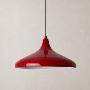 Könnte beinhalten: Eine glänzend rote Pendelleuchte mit einem breiten, glockenförmigen Schirm und einer glatten, reflektierenden Oberfläche. Die Lampe hängt an einem dünnen, strukturierten silbernen Kabel, das vertikal aus der Mitte des kuppelförmigen Metallgehäuses ragt.