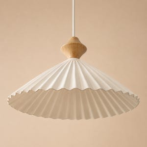 May include: A pendant light fixture featuring a wide, white, pleated conical shade. The top of the shade is accented with a smooth, light-toned wooden finial, and the entire assembly is suspended from a thin, white cord extending upward.