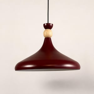 Puede incluir: Una lámpara colgante con una pantalla ancha en forma de campana de color rojo oscuro. La parte superior de la pantalla está decorada con una pequeña pieza circular roja a juego y una esfera de madera de color claro natural, suspendida por un cable negro.
