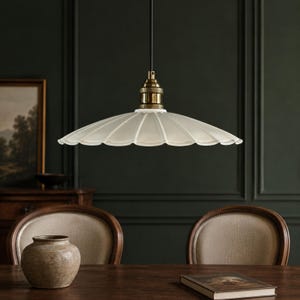 May include: A pendant light fixture featuring a wide, scalloped, frosted white glass shade suspended by a thin black cord. The top of the shade is secured with a textured, metallic brass-toned hardware fitting that connects to the cord.