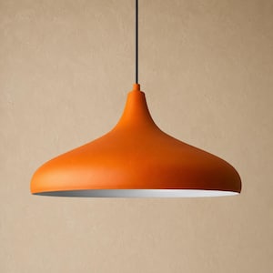 Puede incluir: Una lámpara colgante de color naranja mate con una pantalla ancha en forma de campana. El exterior de la pantalla de metal es de un color naranja sólido y vibrante, mientras que el interior es blanco. Cuelga de un cable negro fino centrado.