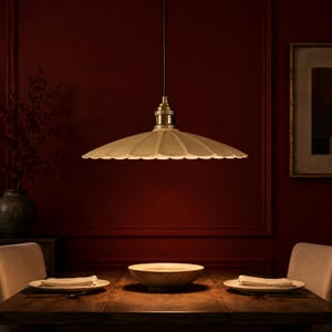 May include: A pendant light fitting featuring a wide, scalloped, off-white shade with a brass-toned metal socket and a black cord. The light is suspended above a wooden dining table set with two plates, napkins, and a central bowl.