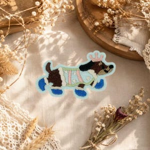 May include: An embroidered patch featuring a brown and black dog wearing a light blue sweater with pink stripes and a pink crown on its head. The dog has blue accents on its paws and tail, with a light blue border outlining the entire shape.