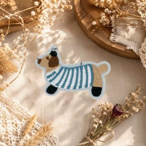Puede incluir: Un parche bordado con forma de perro de perfil. El perro tiene la cabeza de color canela, orejas negras y patas negras. Lleva una camiseta de rayas horizontales azules y blancas. El parche tiene un borde azul claro y tela de base blanca.