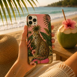 May include: A pink phone case with a leopard design and tropical foliage. The case is held in a hand, with a beach scene in the background, including a coconut drink with a flower.