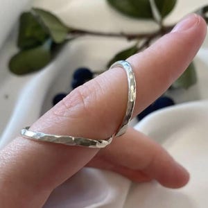 Könnte beinhalten: Zwei silberfarbene Ringe mit einer gehämmerten, strukturierten Oberfläche. Die Ringe haben ein schmales, abgerundetes Banddesign mit einer unregelmäßigen, reflektierenden Oberfläche. Jeder Ring wird an einem Finger getragen und zeigt den metallischen Glanz sowie die handgearbeitete Optik.
