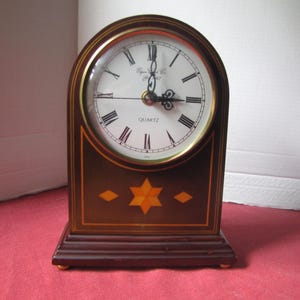 May include: A dark brown wooden mantel clock with an arched top and a white circular face featuring black Roman numerals. The clock displays the text Elgin Clock Co. and QUARTZ. A six-pointed star inlay in orange and yellow sits below the clock face.