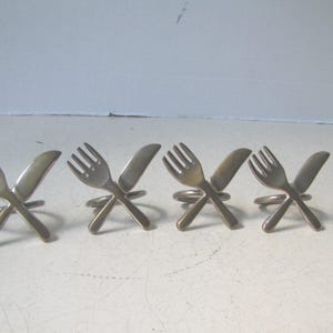 May include: A set of four metallic napkin rings, each featuring a decorative design of a fork and a knife crossed in an X shape. The napkin rings have a smooth, polished finish and a circular base for holding napkins securely in place.