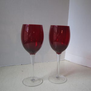 May include: Two red wine glasses with clear stems and bases. The bowls of the glasses are a deep, rich red color, while the stems and bases are transparent. The glasses are positioned side by side.