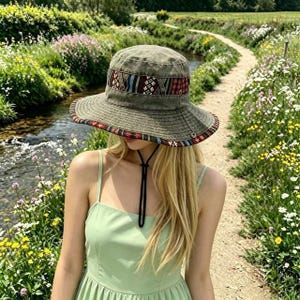May include: Olive green bucket hat with a patterned band in red, black, and white. The hat has a wide brim and a black chin strap. The hat is worn by a person wearing a light green dress, standing on a path next to a stream.