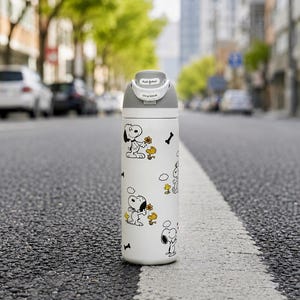 Puede incluir: Una botella de agua aislada blanca con ilustraciones de Snoopy y Woodstock. La botella tiene una tapa gris con un mecanismo de bot&oacute;n y el texto "Push Button" y "Slide to Unlock". El dise&ntilde;o incluye gr&aacute;ficos de huesos y flores negros.
