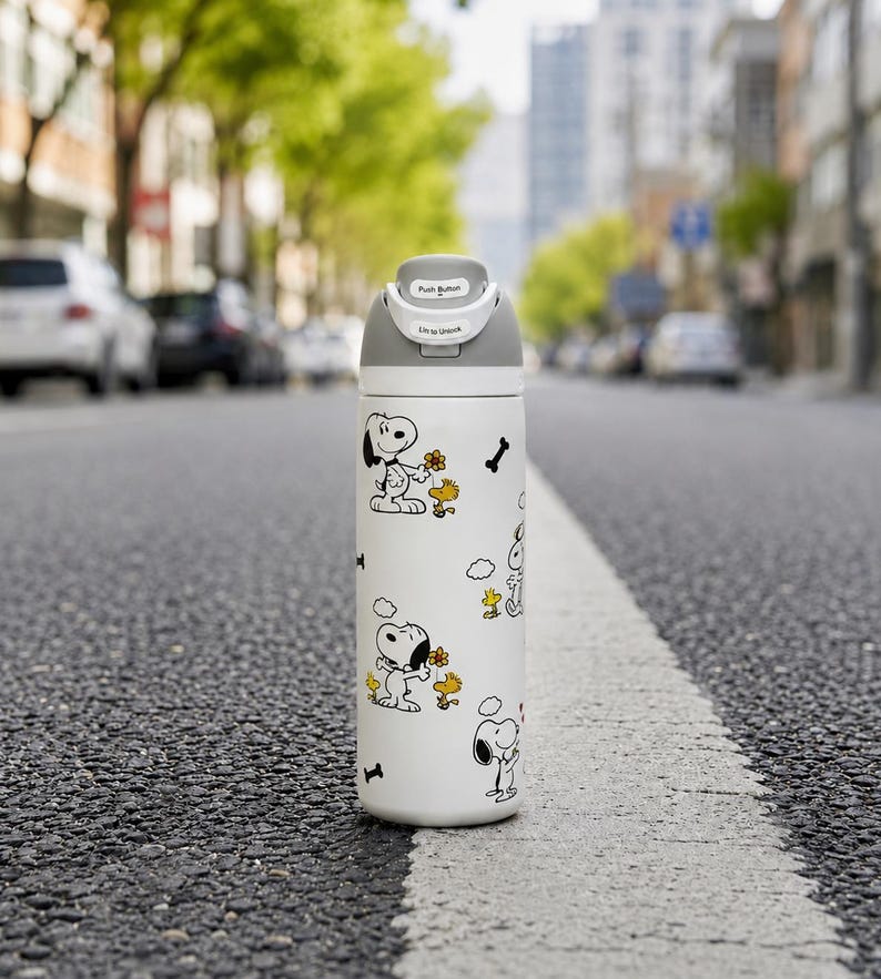 Puede incluir: Una botella de agua aislada blanca con ilustraciones de Snoopy y Woodstock. La botella tiene una tapa gris con un mecanismo de bot&oacute;n y el texto "Push Button" y "Slide to Unlock". El dise&ntilde;o incluye gr&aacute;ficos de huesos y flores negros.