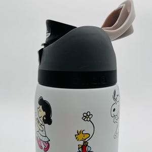 May include: White water bottle featuring cartoon characters. The bottle has a black lid and a light pink flip-top. The bottle is decorated with colorful images of cartoon characters, including Snoopy and Woodstock.