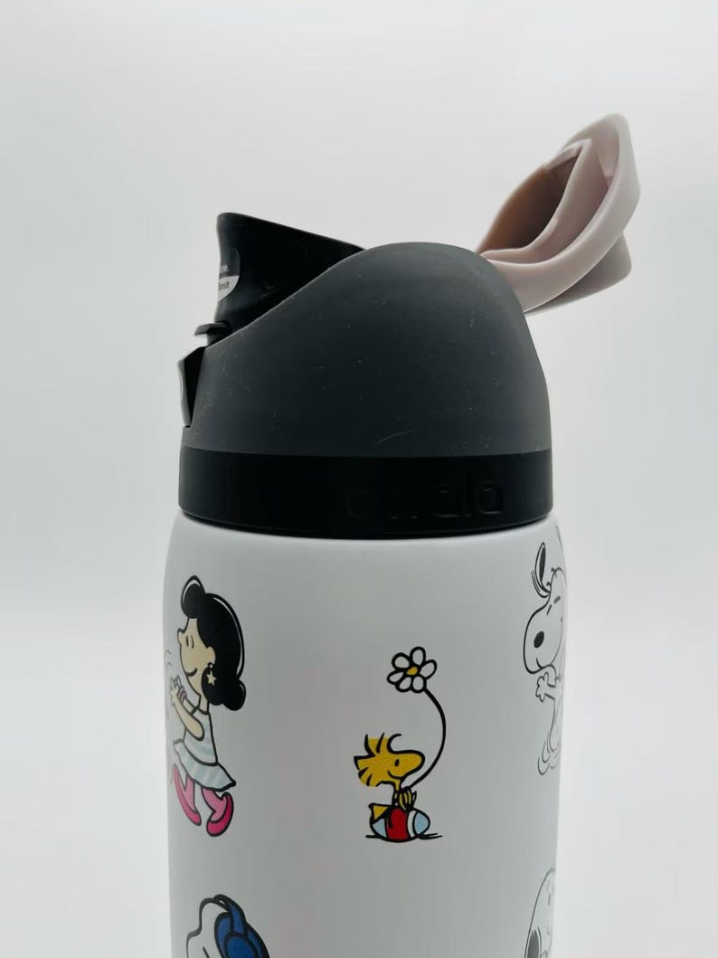 May include: White water bottle featuring cartoon characters. The bottle has a black lid and a light pink flip-top. The bottle is decorated with colorful images of cartoon characters, including Snoopy and Woodstock.