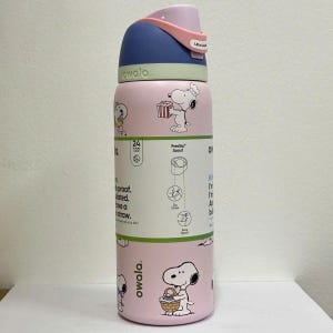 May include: A pink Owala FreeSip water bottle featuring Snoopy illustrations. The bottle has a blue and pink lid with a carrying handle. It is insulated and has a straw. The bottle is designed to keep drinks cold for 24 hours.